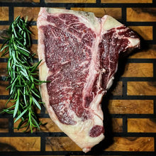Load image into Gallery viewer, A raw Angus T-bone steak on a wooden surface with a sprig of rosemary to the side.