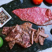 Cargar imagen en el visor de la galería, A display of wagyu carne asada, with slices of beef, a whole beefsteak, and seasonings like salt and pepper, presented on a dark stone surface with tomatoes and a lime in the background.